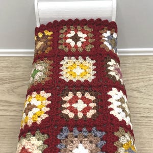 May include: A miniature bed with a handmade crochet blanket. The blanket features a patchwork design with squares in shades of red, brown, yellow, and cream. The bed frame is white.