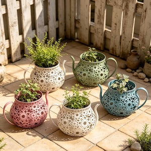 May include: Five metal teapot-shaped planters with intricate floral cutouts, each containing a small green plant. The planters are finished in white, light green, pink, cream, and blue, featuring a curved handle and a spout on opposite sides.