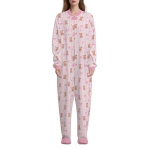 May include: A pink and white striped onesie featuring a teddy bear and bow design. The long-sleeved jumpsuit has a front zip, pink cuffs, and pink trim around the collar and feet.