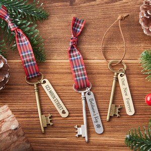 May include: Three decorative keys with engraved tags and plaid ribbons. One tag reads "123 Main Street" with a heart, another says "Baldwin's Key" with a snowflake, and the last says "Our New Forever". The keys are on a wooden surface.