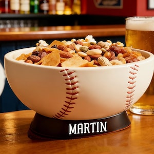 Puede incluir: Un cuenco de cerámica blanco con forma de béisbol, lleno de frutos secos y aperitivos. El cuenco tiene costuras rojas y una base negra con el nombre "MARTIN". Una jarra de cerveza está al lado del cuenco.