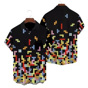 Op de afbeelding: Zwart overhemd met korte mouwen en een kleurrijk pixelpatroon. Het patroon toont vierkanten in verschillende kleuren, die doen denken aan een klassiek videospel. Het overhemd heeft een borstzak en wordt op een hanger getoond.