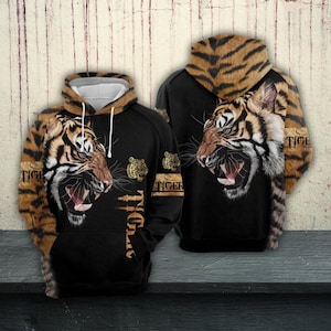 Può includere: Felpa con cappuccio nera con un design dettagliato di volto di tigre, bocca aperta e denti visibili. Il cappuccio e le maniche hanno un motivo a strisce di tigre. La parola "TIGER" è stampata sulle maniche e sul lato. La felpa con cappuccio è esposta su una superficie di legno.