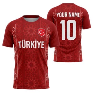 Könnte beinhalten: Ein weinrotes Fußballtrikot mit weißen Akzenten und einem geometrischen Muster. Auf der Vorderseite steht das Wort "TÜRKIYE" und ein türkisches Flaggenemblem. Auf der Rückseite stehen die Nummer 10 und der Text "YOUR NAME".