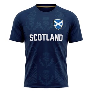 May include: Navy blue t-shirt with the word "SCOTLAND" in white block letters. The shirt features a thistle pattern and the Scottish flag on the upper chest. The shirt has short sleeves and a crew neck.