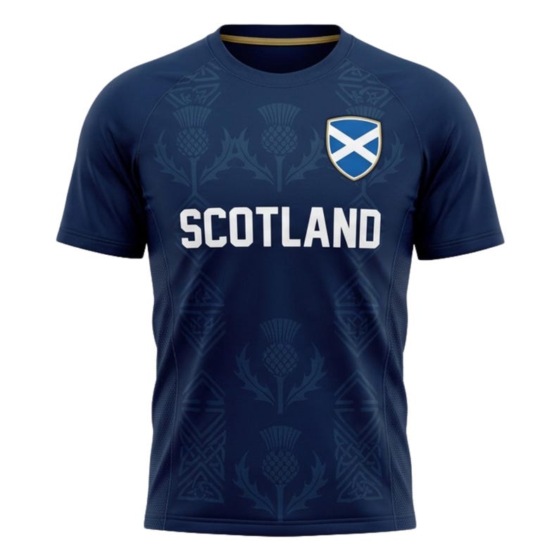 May include: Navy blue t-shirt with the word "SCOTLAND" in white block letters. The shirt features a thistle pattern and the Scottish flag on the upper chest. The shirt has short sleeves and a crew neck.