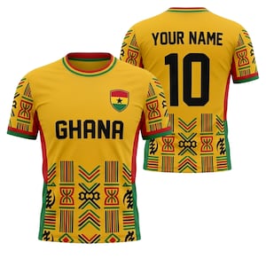 Puede incluir: Una camiseta de fútbol amarilla de Ghana con detalles rojos y verdes. La parte delantera muestra la palabra "GHANA" y un emblema nacional. La parte trasera presenta el número "10" en negro y el texto "YOUR NAME". La camiseta tiene un diseño.