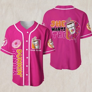 May include: A pink baseball jersey with white trim and buttons. The front features a donut graphic and the words "Dunkin' Donuts". The back displays the phrase "She Wants The" with a coffee cup and donut graphic.