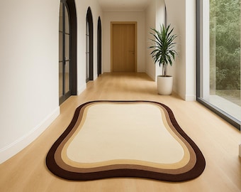 May include: A beige, brown, and tan area rug with an organic shape. The rug is placed on a light wood floor in a hallway. A tall potted plant and a wooden door are visible in the background.