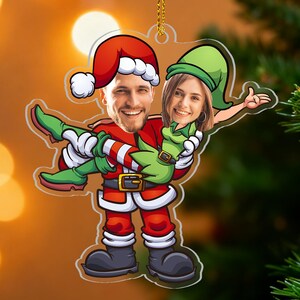 Può includere: Un ornamento natalizio raffigurante una figura di Babbo Natale che trasporta un elfo. L'ornamento ha uno sfondo trasparente con i volti di due persone. La figura di Babbo Natale indossa un abito e un cappello rossi, e l'elfo indossa un abito e un cappello verdi.
