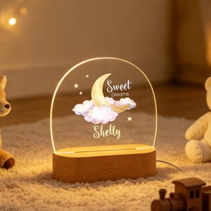 May include: A semi-circular acrylic night light with a wooden base, featuring a printed design of a crescent moon, pink clouds, and stars. The text "Sweet Dreams" and the name "Shelly" are printed in white script on the clear acrylic surface.