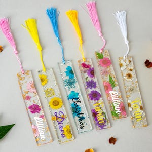 May include: A selection of clear resin bookmarks, each with dried flowers and gold leaf inclusions. The bookmarks have colourful tassels and feature names or the word "Christmas". The rectangular bookmarks have a floral design.