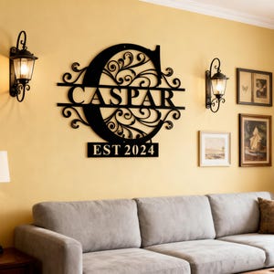 May include: Black metal wall art featuring the name "CASPAR" within a decorative "C" initial, with "EST 2024" below. The art is mounted on a yellow wall, flanked by black sconces and framed artwork, above a gray sofa.