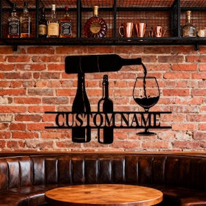 May include: Black metal wall art depicting a wine bottle pouring into a wine glass, with two wine bottles to the left. The text "CUSTOM NAME" is centered below the bottles and glass. The art is mounted on a brick wall.