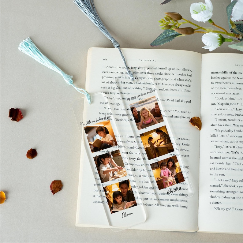 May include: A clear acrylic bookmark with a tassel, featuring a series of small photos. The photos show a young girl in various settings. The bookmark is placed in an open book with text.