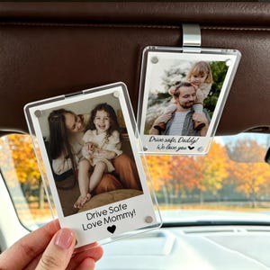 May include: Two clear acrylic car visor photo holders with family photos. One holder says "Drive Safe Love Mommy!" and the other says "Drive safe, Daddy! We love you". The holders are attached to a car visor.