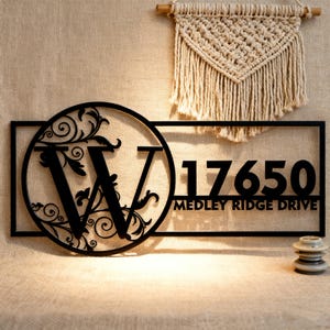 Könnte beinhalten: Schwarzes Metall-Hausnummernschild mit dem Buchstaben "W" in einem kreisförmigen Design mit floralen Akzenten. Das Schild zeigt die Zahlen "17650" und "MEDLEY RIDGE DRIVE". Ein Makramee-Wandbehang befindet sich im Hintergrund.