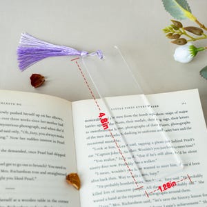 May include: A clear, rectangular bookmark with a purple tassel, measuring 4.8 inches long and 1.28 inches wide. The bookmark is placed on an open book. The tassel is made of multiple strands of purple thread.