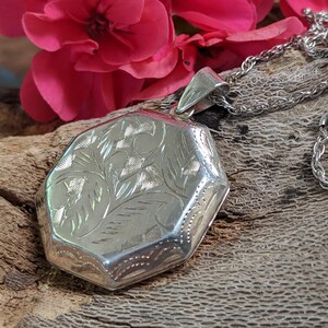 May include: A silver locket with an intricate floral design hangs from a silver chain. The locket is octagonal in shape and has a smooth, polished finish.