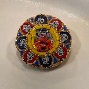 May include: A round, colorful mosaic brooch with a floral design in the center. The brooch is made of small, colorful glass tiles and is surrounded by a gold-colored metal frame.