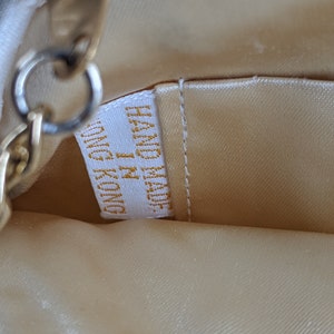 Puede incluir: Primer plano del forro de un bolso de tela beige con una cadena dorada y una etiqueta blanca que dice "Made in China" en letras doradas.