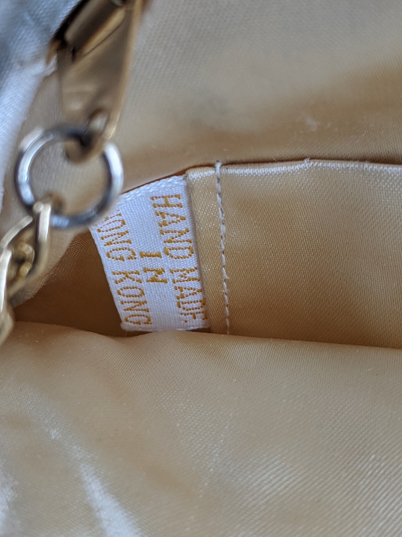Puede incluir: Primer plano del forro de un bolso de tela beige con una cadena dorada y una etiqueta blanca que dice "Made in China" en letras doradas.