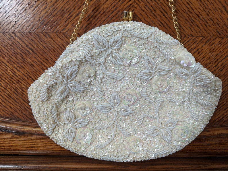 Puede incluir: Un bolso de noche blanco de cuentas con una correa de cadena dorada. El bolso est&aacute; cubierto de lentejuelas y cuentas blancas, formando un patr&oacute;n floral.