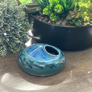 May include: A small, round, blue and green ceramic planter with a hole in the top. The planter has a white glaze and a black design.