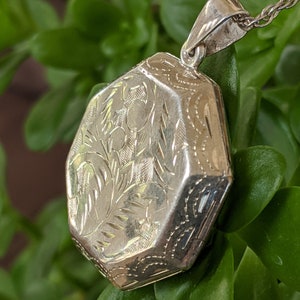 May include: A silver locket with an octagonal shape and intricate floral engravings. The locket is attached to a silver chain.