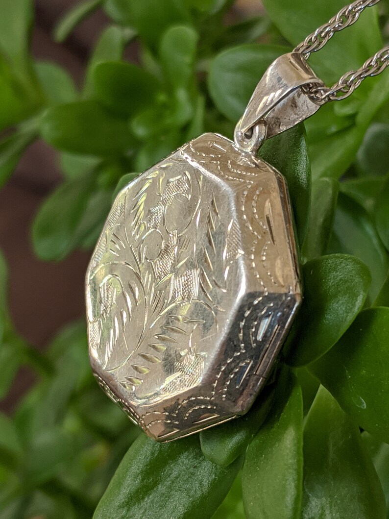 May include: A silver locket with an octagonal shape and intricate floral engravings. The locket is attached to a silver chain.