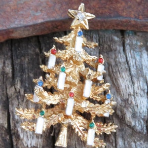 May include: A gold-toned Christmas tree brooch with white candle accents and colorful gems. The tree is topped with a star featuring a clear gem. The brooch is set against a rustic wooden background.