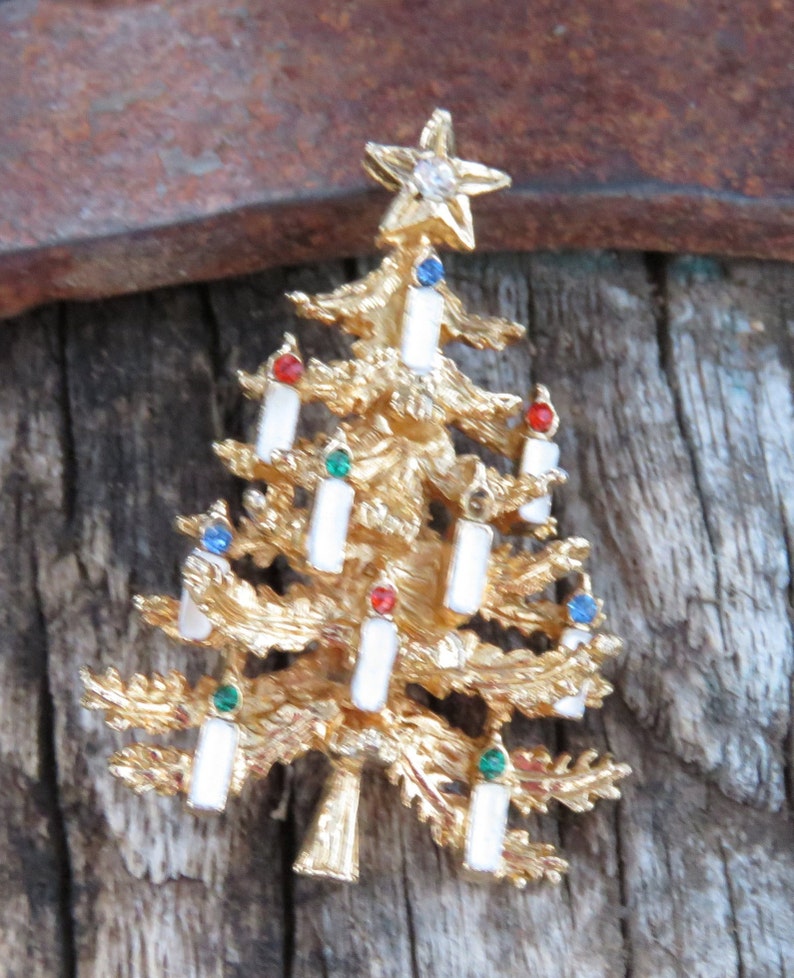 May include: A gold-toned Christmas tree brooch with white candle accents and colorful gems. The tree is topped with a star featuring a clear gem. The brooch is set against a rustic wooden background.