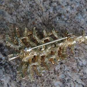 May include: A gold-colored Christmas tree brooch with a star on top. The back of the brooch features a pin for attachment. The brooch is made of metal and has a textured surface, with a detailed design.
