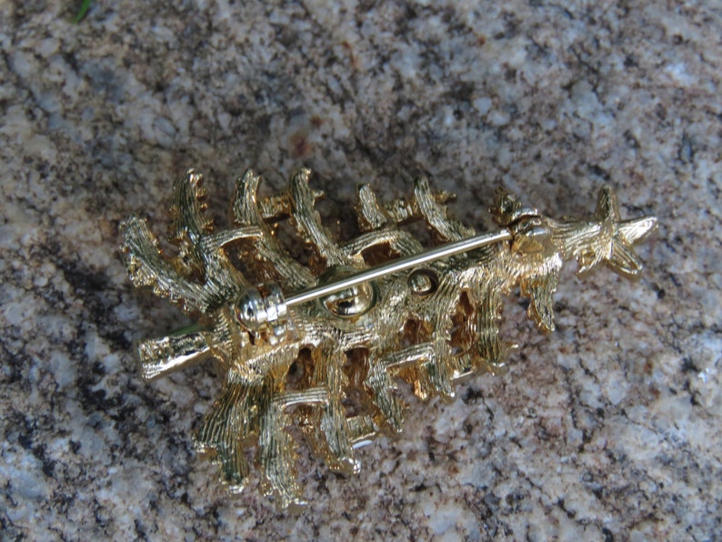May include: A gold-colored Christmas tree brooch with a star on top. The back of the brooch features a pin for attachment. The brooch is made of metal and has a textured surface, with a detailed design.