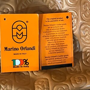 Pode incluir: Etiqueta laranja e branca com o texto "Marino Orlandi" e "100% Feito na It&aacute;lia". A etiqueta tamb&eacute;m apresenta um logotipo com um "M" estilizado. O texto "O produto original &eacute; a garantia de venda." est&aacute; em uma etiqueta laranja menor com texto preto.