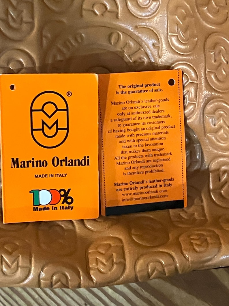 Pode incluir: Etiqueta laranja e branca com o texto "Marino Orlandi" e "100% Feito na It&aacute;lia". A etiqueta tamb&eacute;m apresenta um logotipo com um "M" estilizado. O texto "O produto original &eacute; a garantia de venda." est&aacute; em uma etiqueta laranja menor com texto preto.