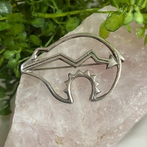 May include: A silver-toned metal brooch with a mountain and sun design. The brooch features a stylized mountain range with arrows and a sun-like shape. The brooch is set against a pink stone surface with green foliage in the background.