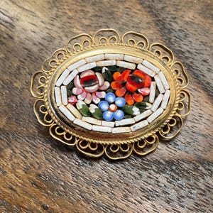 May include: A gold oval-shaped brooch with a floral mosaic design. The mosaic features red, orange, blue, green, and white tiles.