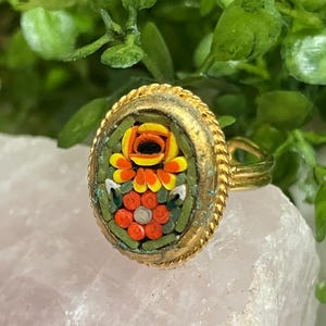 May include: Gold-toned ring with an oval mosaic design featuring a floral pattern in shades of orange, yellow, and red, set against a green background. The ring has a textured, rope-like border.