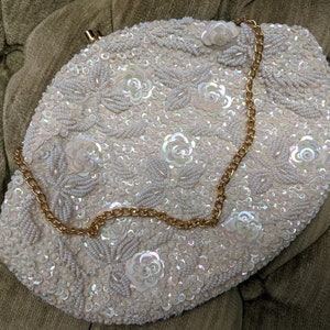 Puede incluir: Un bolso de mano blanco de cuentas con una cadena dorada. El bolso est&aacute; decorado con lentejuelas iridiscentes y cuentas de imitaci&oacute;n de perlas en un patr&oacute;n floral.