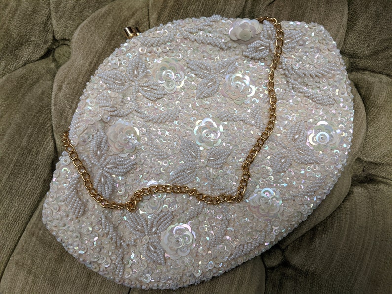 Puede incluir: Un bolso de mano blanco de cuentas con una cadena dorada. El bolso est&aacute; decorado con lentejuelas iridiscentes y cuentas de imitaci&oacute;n de perlas en un patr&oacute;n floral.