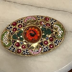 May include: An oval-shaped mosaic brooch with a gold frame. The center of the brooch features a red flower with a black center. The mosaic is made up of small, colorful tiles in a variety of patterns.