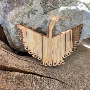 May include: A carved wooden bird with black dots around the edges. The bird is in flight with its wings spread out. The wood is a light brown color with darker brown stripes.