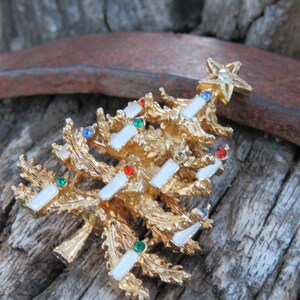 May include: A gold-toned Christmas tree brooch with a textured surface. The tree is adorned with white rectangular accents, and colorful gems. A star tops the tree, adding a festive touch to this holiday-themed jewelry.