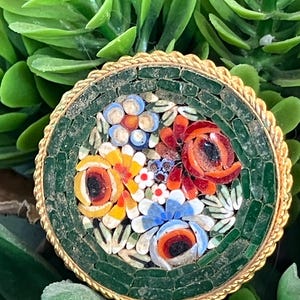 May include: A round, green mosaic brooch with a gold rim. The mosaic features a floral design with red, orange, yellow, blue, and white flowers.