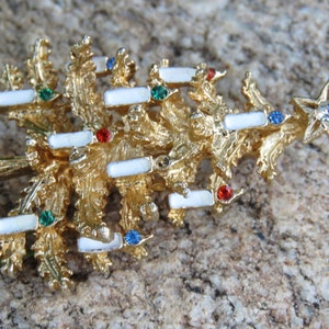 May include: A gold-toned Christmas tree brooch with white enamel accents and colorful gemstone embellishments. The brooch features a star at the top with a clear rhinestone. A festive accessory for the holiday season.