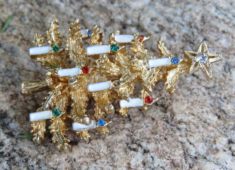 May include: A gold-toned Christmas tree brooch with white enamel accents and colorful gemstone embellishments. The brooch features a star at the top with a clear rhinestone. A festive accessory for the holiday season.