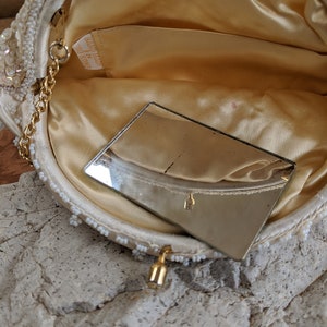 Puede incluir: Un bolso de noche blanco con cuentas, con una cadena dorada y un peque&ntilde;o espejo en el interior. El bolso tiene una etiqueta que dice "Hand Made in Hong Kong".