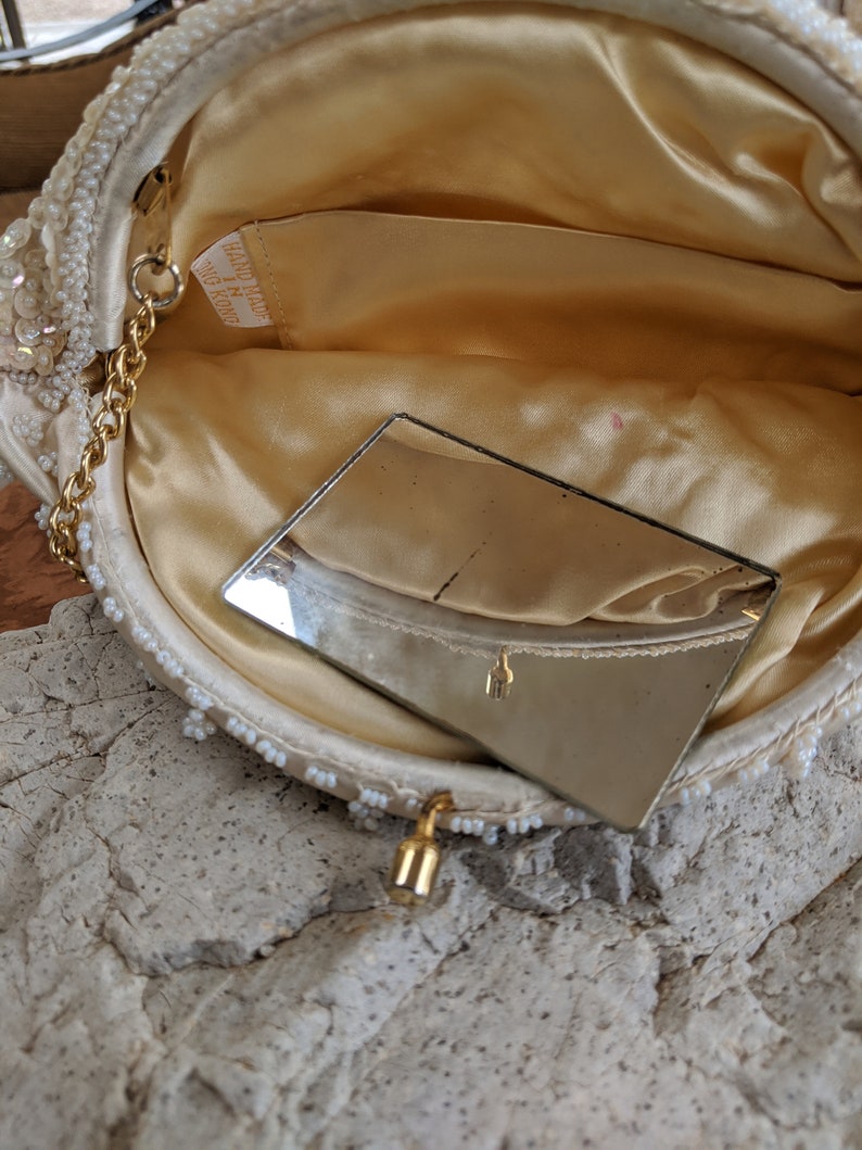 Puede incluir: Un bolso de noche blanco con cuentas, con una cadena dorada y un peque&ntilde;o espejo en el interior. El bolso tiene una etiqueta que dice "Hand Made in Hong Kong".