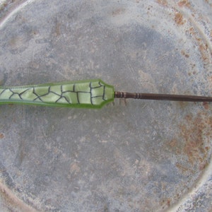 May include: A vintage crochet hook with a light green, mosaic-patterned handle and a dark metal hook. The handle has a geometric design with black lines. The hook is made of metal and is curved at the end.